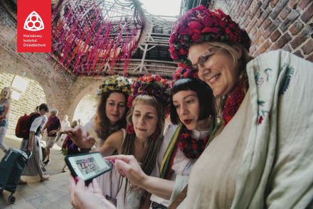 Cztery kobiety w wiankach na głowie oglądają zdjęcie, które zrobiły telefonem pająkowi. Ozdoba wisi nad nimi w Bramie Krakowskiej.