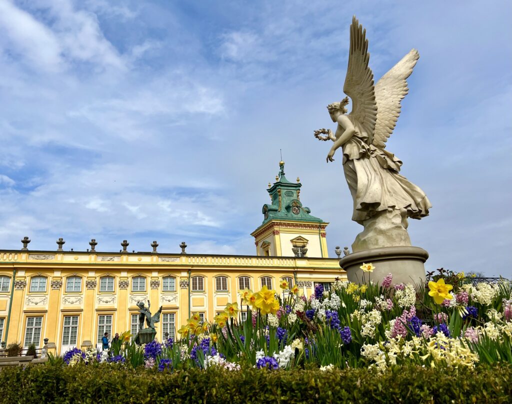 Na pierwszym planie widok rzeźby przestawiającej kobietę-anioła . W tle fragment Pałacu Króla Jana II w Wilanowie