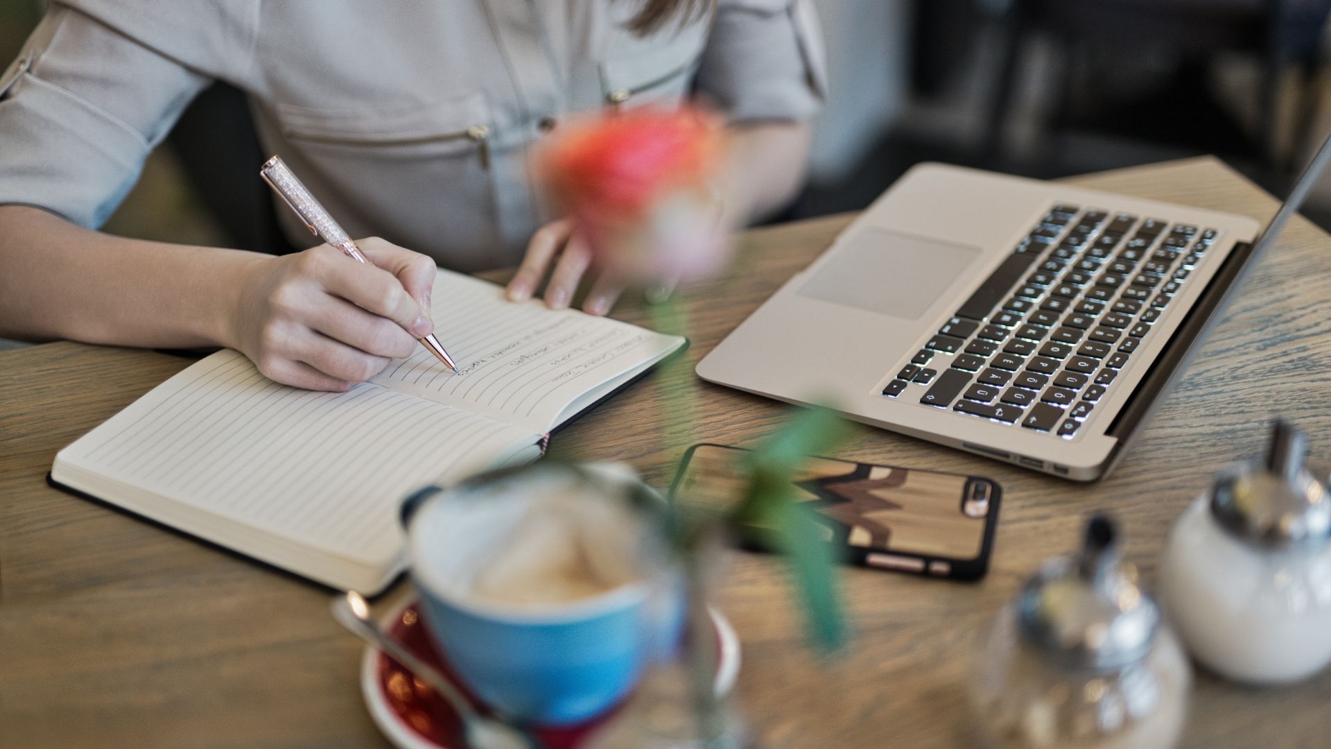 Kobieta siedząca przy biurku pisze w notatniku. Na biurku leży otwarty laptop, telefon i niebieska filiżanka, róża