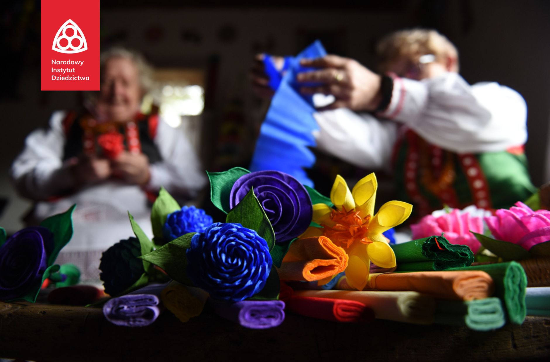 Dwie kobiety pracują nad kolorowymi papierowymi rękodziełami, Na pierwszym planie papierowe kwiaty i dekoracje.