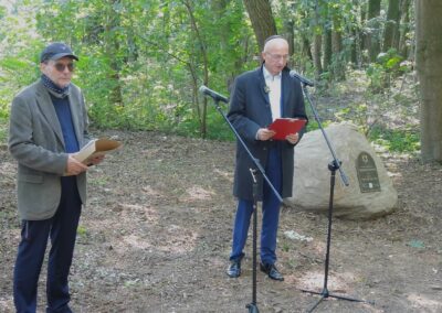 Dwóch mężczyzn stojących przed mikrofonem. W tle kamień z tablicą w języku polskim i hebrajskim